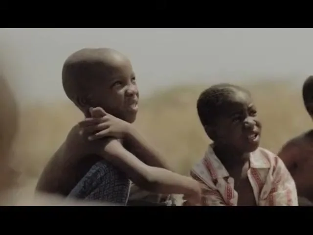 Two young African boys, one shirtless and one in a patterned shirt, sit outdoors looking up with expressions of awe and wonder.