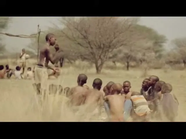 A young boy, shirtless and wearing light shorts, stands and gestures to a group of children sitting in a circle in a dry, grassy field.