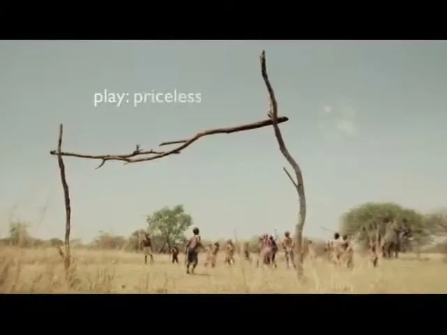 A group of children in traditional attire play soccer with a makeshift goalpost in a dry, grassy field under a clear sky.
