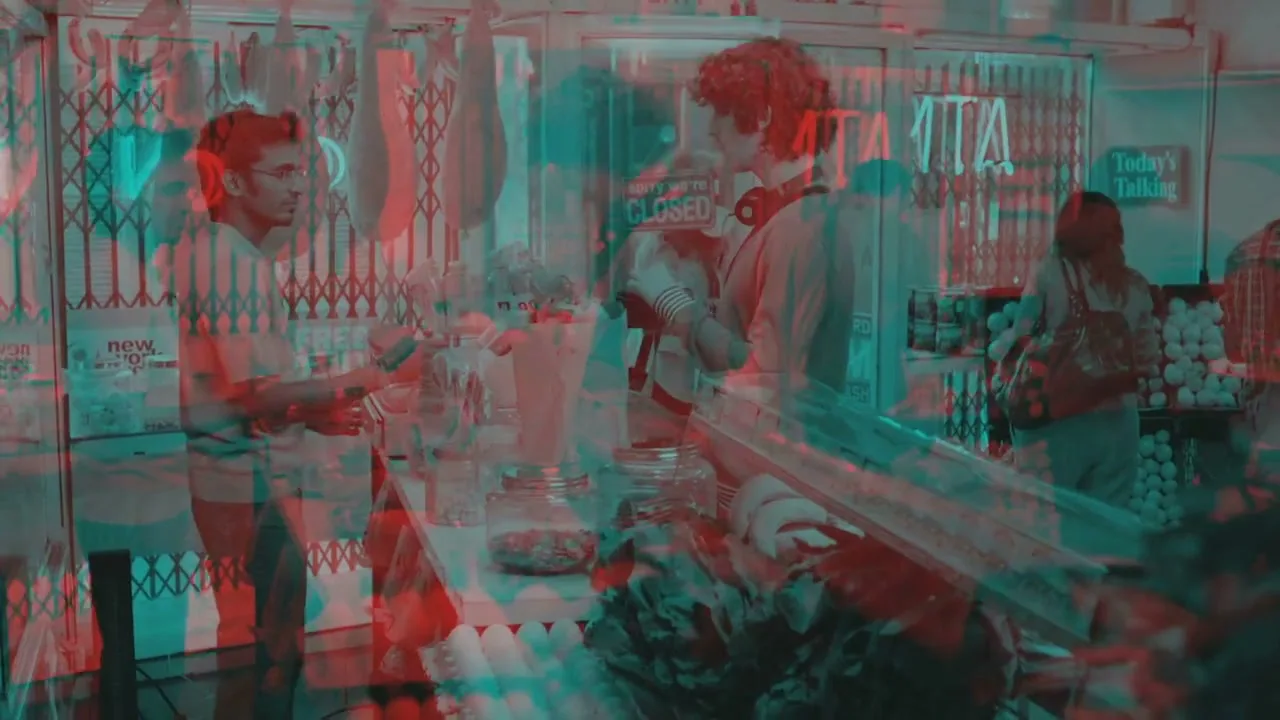 A young man with curly hair and headphones around his neck stands at a counter in a busy market, interacting with a vendor.