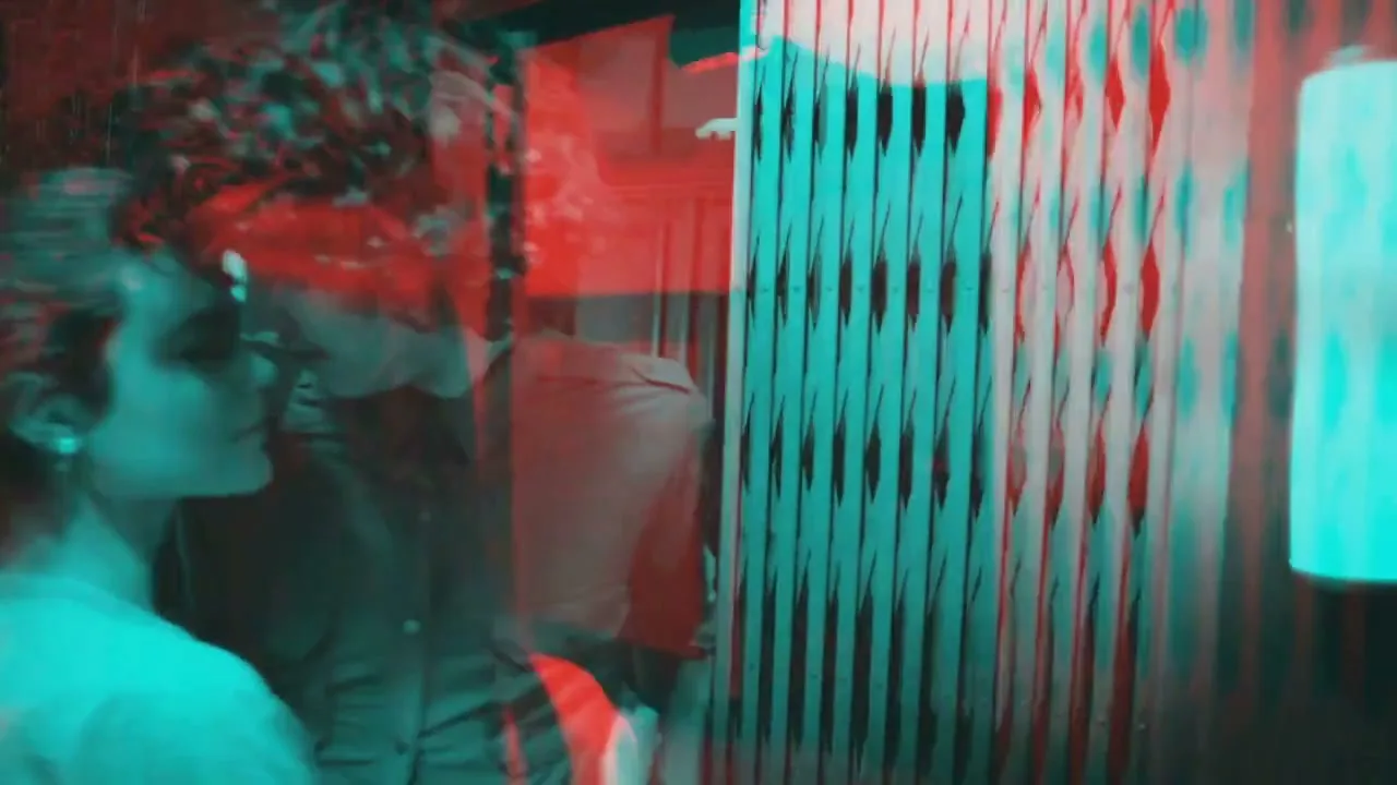 A young woman with dark hair and a man with curly hair are seen in a split-color, 3D-like effect, kissing passionately in a dimly lit, industrial-looking hallway.