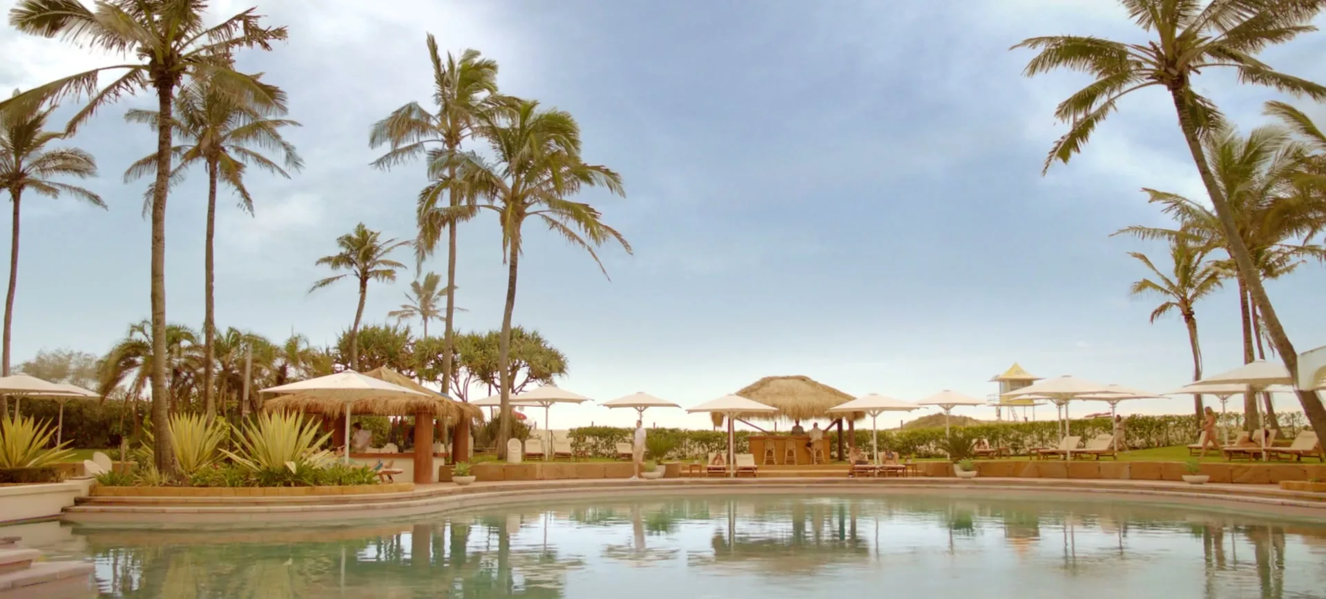 A wide shot of a luxurious resort pool area with palm trees, umbrellas, and a bar, under a bright sky.