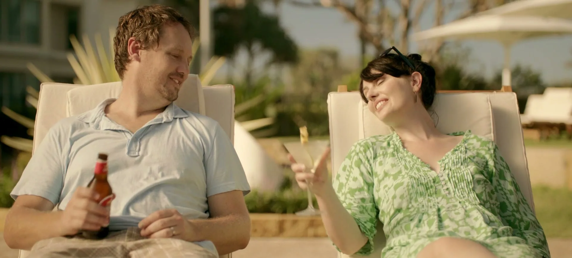 A man and a woman relax in lounge chairs by a pool, smiling at each other, with the man holding a beer and the woman a cocktail.