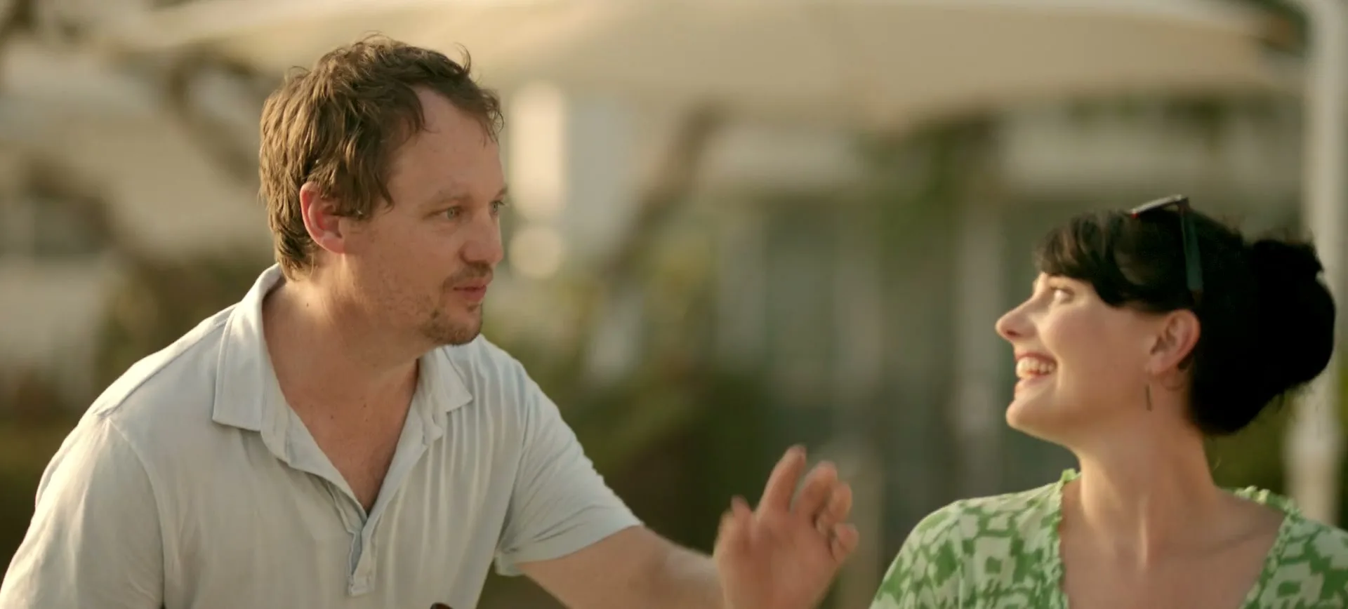 A man in a light blue polo shirt holds a beer bottle and talks to a smiling woman in a green dress at an outdoor gathering during the day.