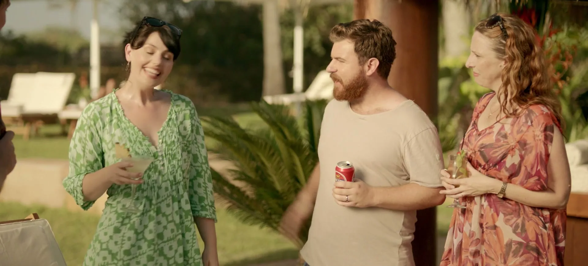 A man with a beard holds a beer can and gestures while talking to two women holding cocktails at an outdoor party.