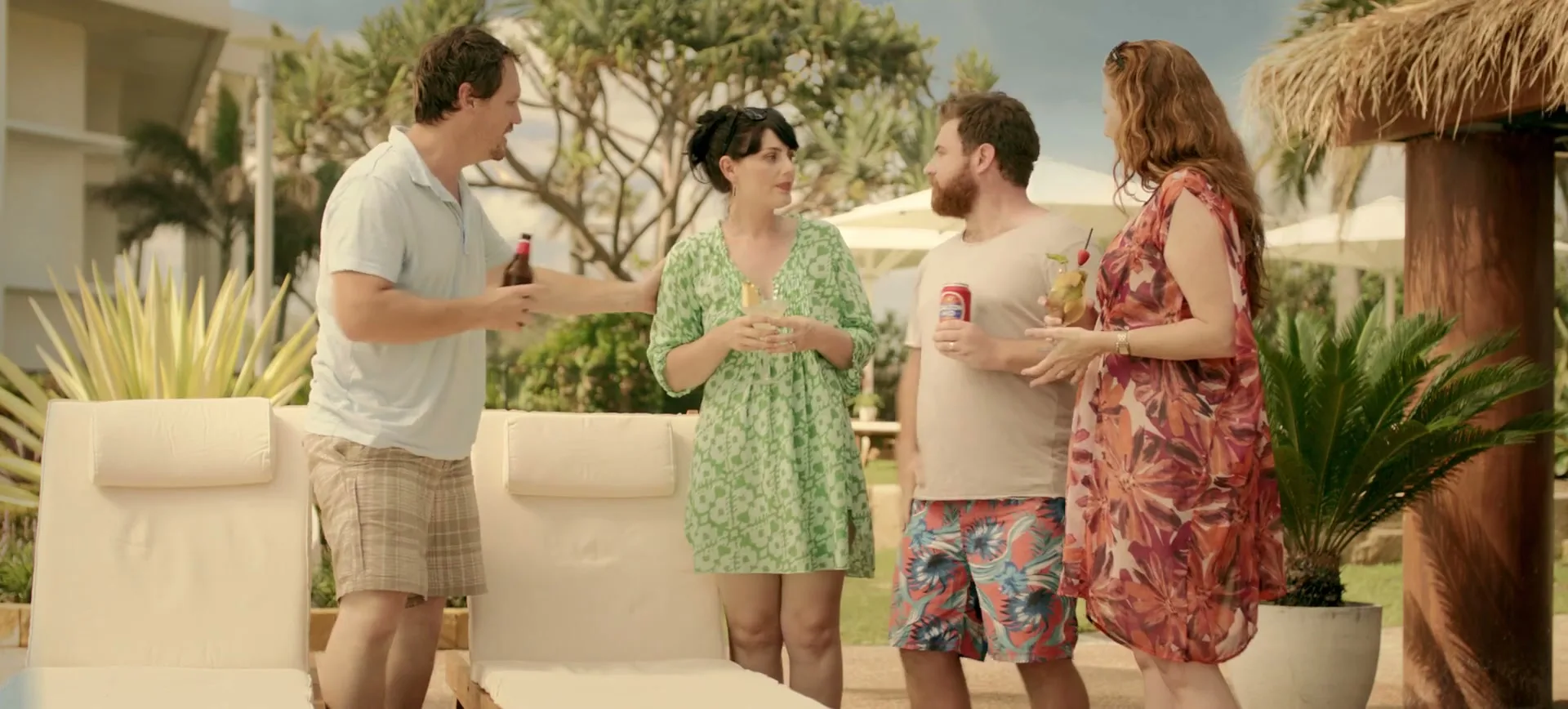 Four friends, two men and two women, stand by a pool, holding drinks and talking, with palm trees and lounge chairs in the background.