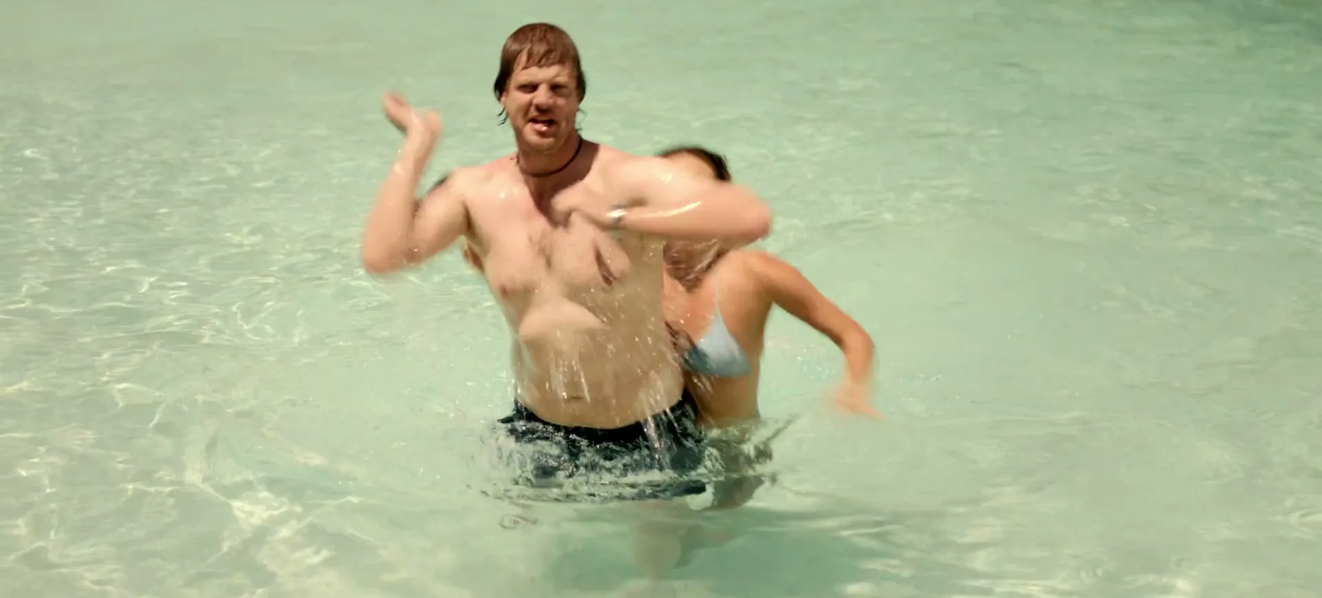 A man and woman are swimming in clear, shallow water, with the woman holding onto the man's back before he flails his arms and pushes her away.