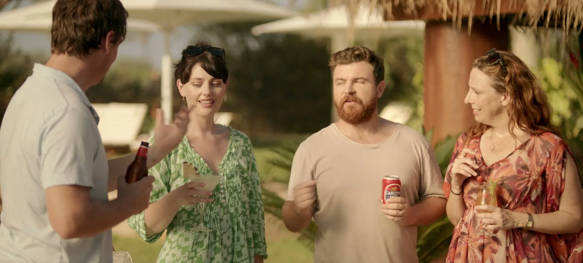 Four friends, two men and two women, are enjoying drinks and conversation outdoors at a sunny resort.