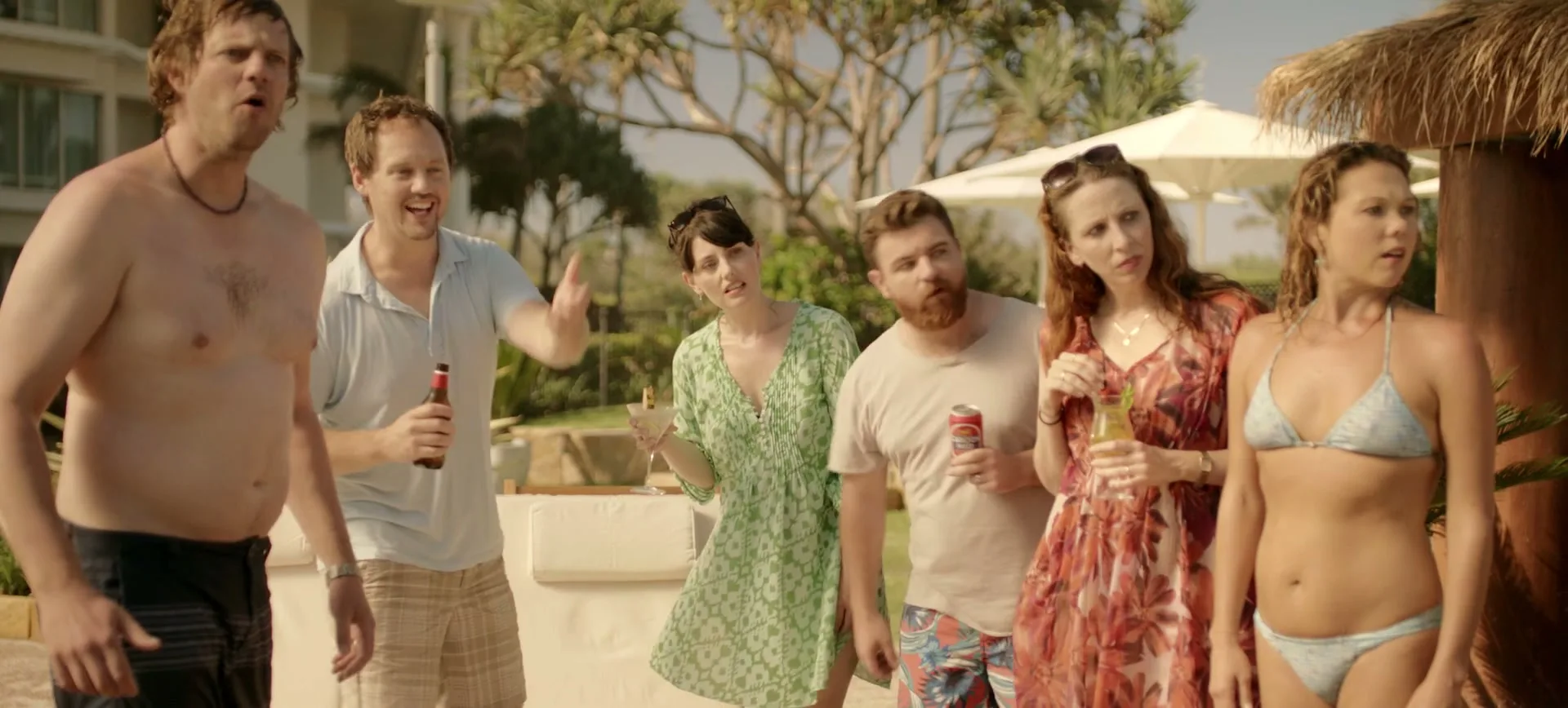 A group of six friends, three men and three women, stand by a pool at a resort, looking surprised and pointing at something off-camera.