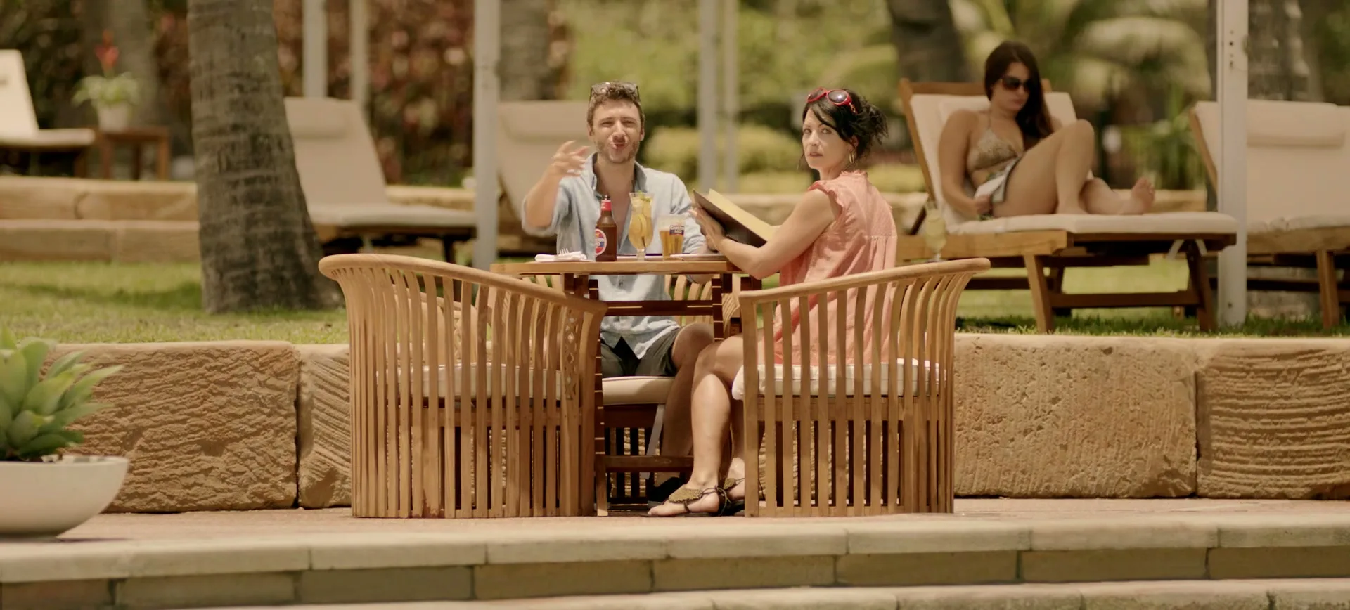 A man and woman sit at a table by a pool, while another woman sunbathes in the background at a tropical resort.