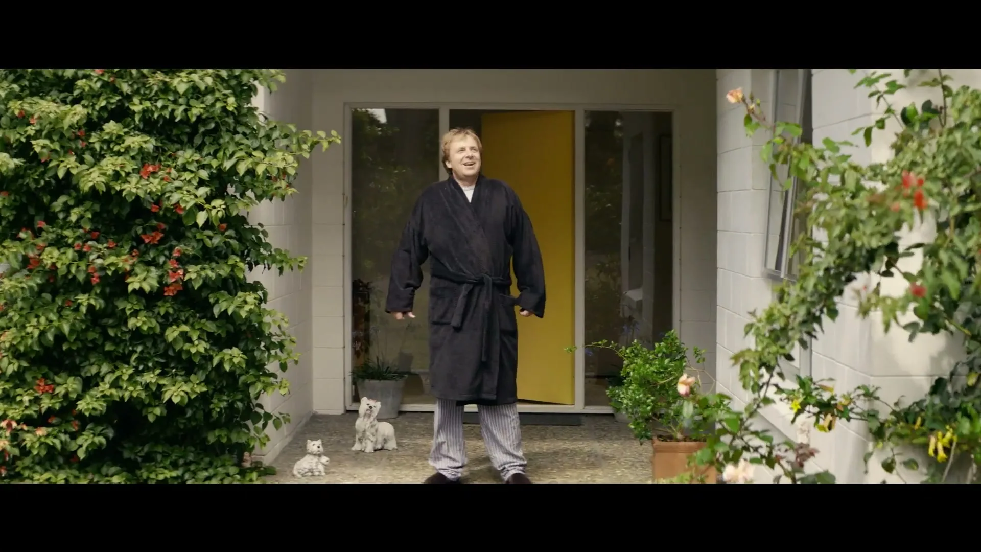 A middle-aged man in a black robe and pajamas steps out of his house, stretches, and then walks back inside, with two small white dog statues on the porch.