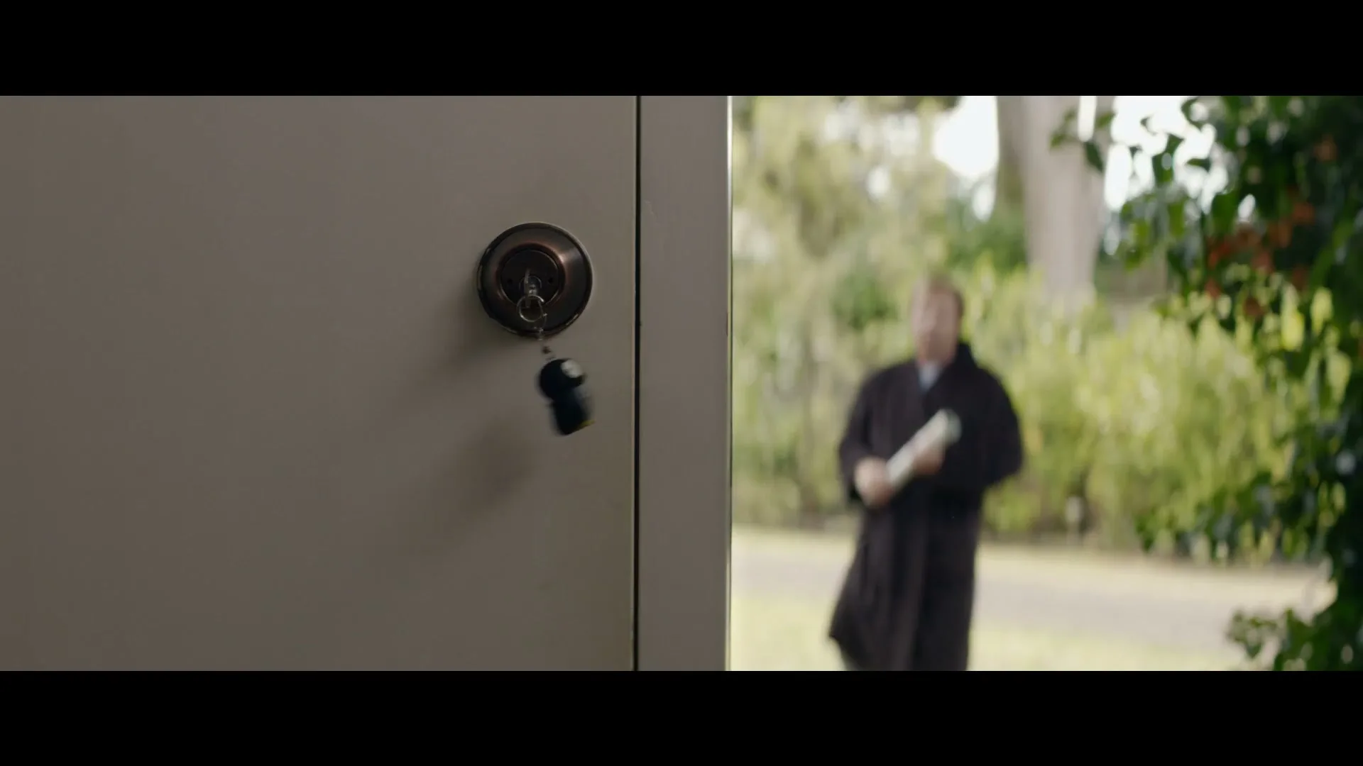 A key dangles from a lock on a light grey door, while a blurry man in a dark robe walks in the background.