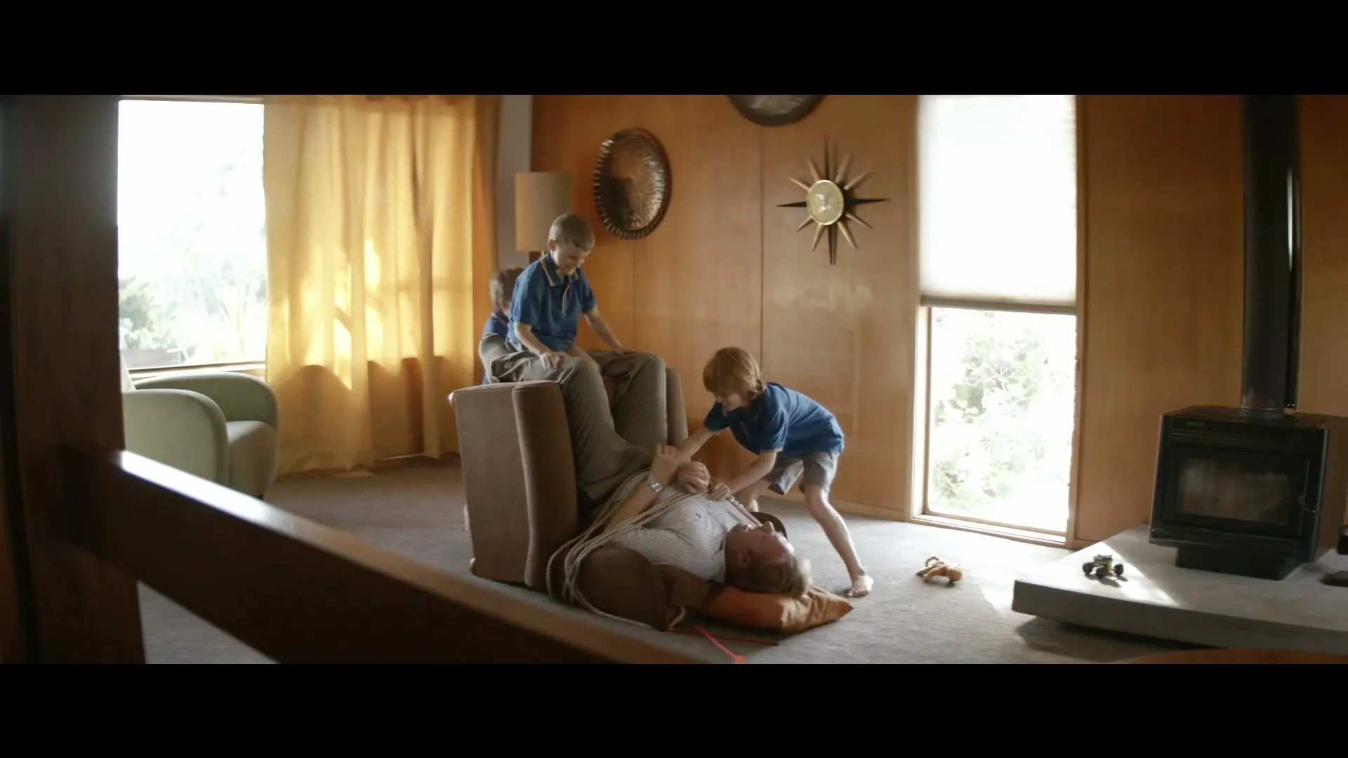 Three young boys in blue shirts play around a man tied to a recliner in a mid-century modern living room.