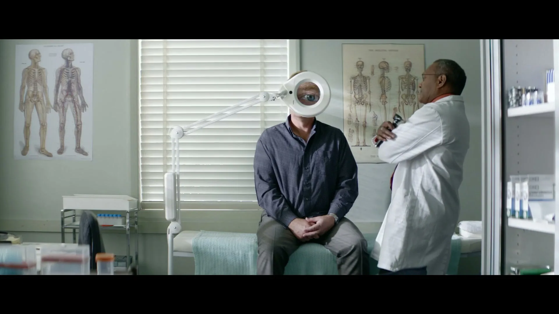 A male patient sits on an examination table with a magnifying lamp in front of his face, while a male doctor stands beside him in a doctor's office.