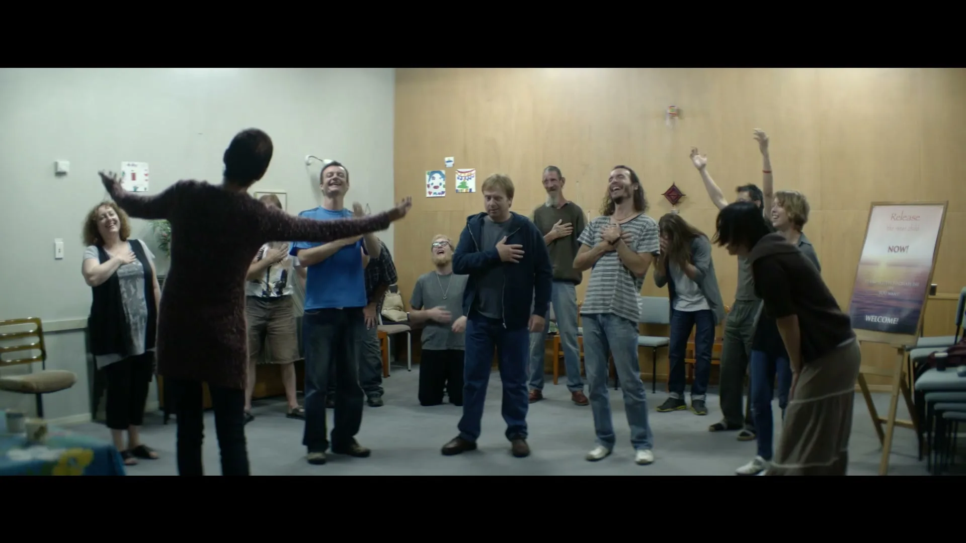 A diverse group of adults in casual clothes participate in a group activity, raising their hands and laughing in a brightly lit community hall.