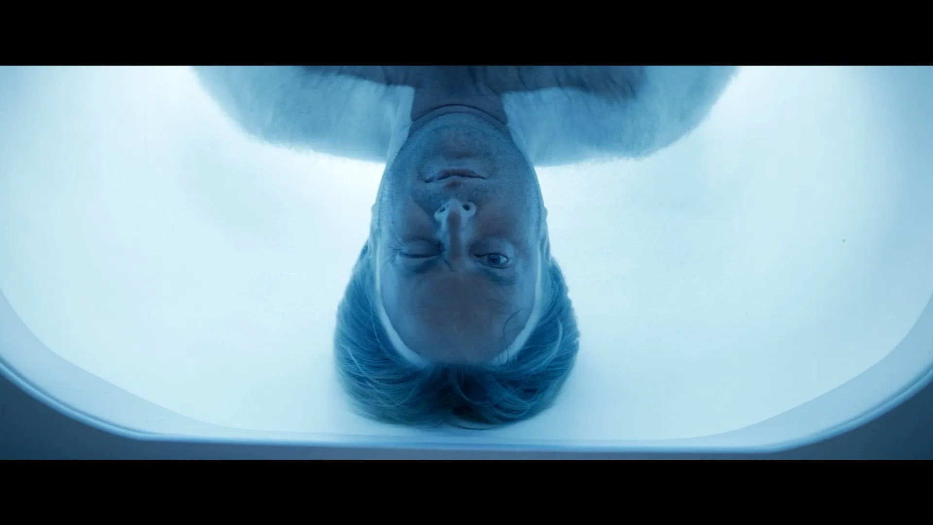 A man with blonde hair and a beard is seen upside down, floating in a blue-lit tank, with his eyes open.