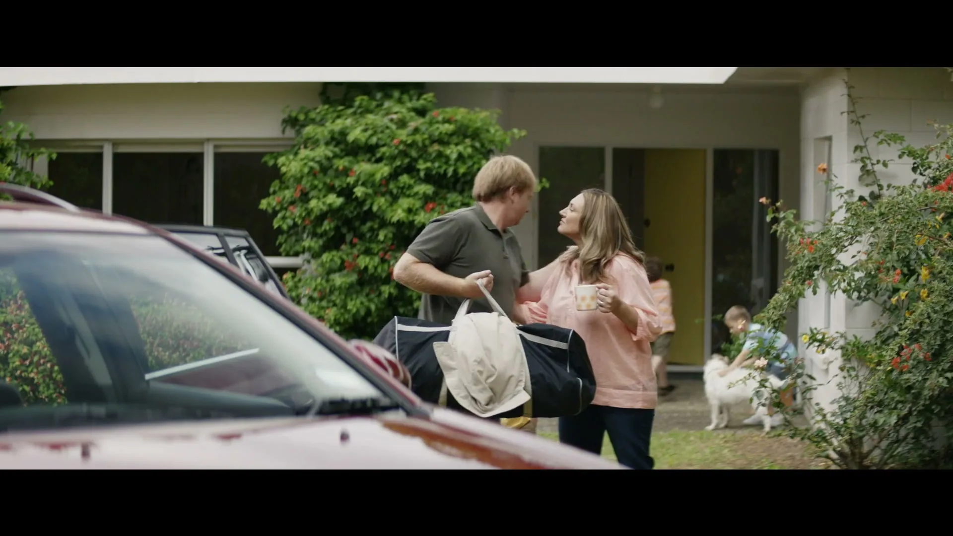 A man and woman stand outside a house, talking and kissing, while children and a dog are visible in the background.