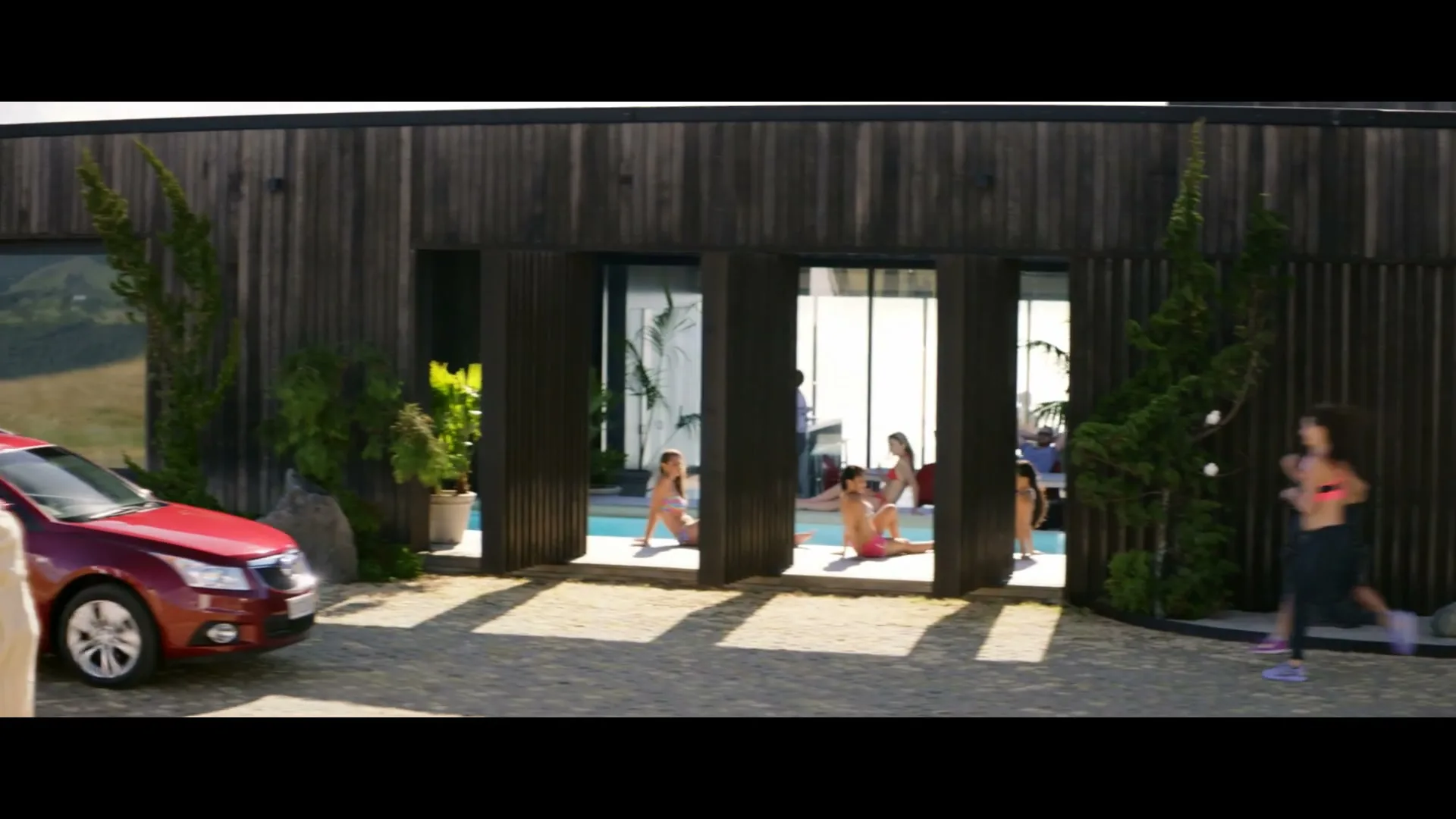 A red Holden car drives past a modern house with people lounging by a pool, while a man practices tai chi in the foreground.