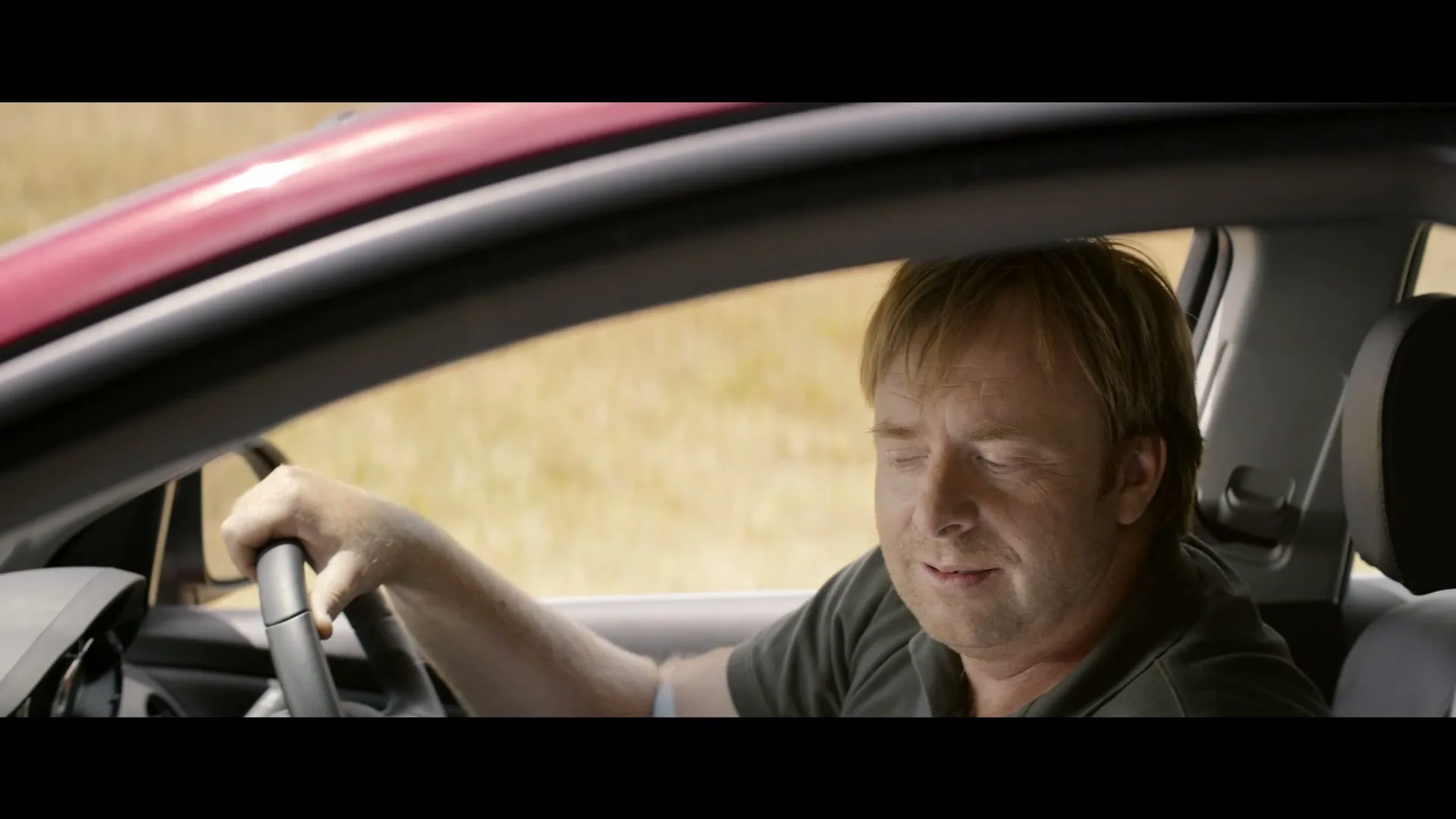 A man with blonde hair and a dark shirt drives a red car, looking around with a slightly confused expression.