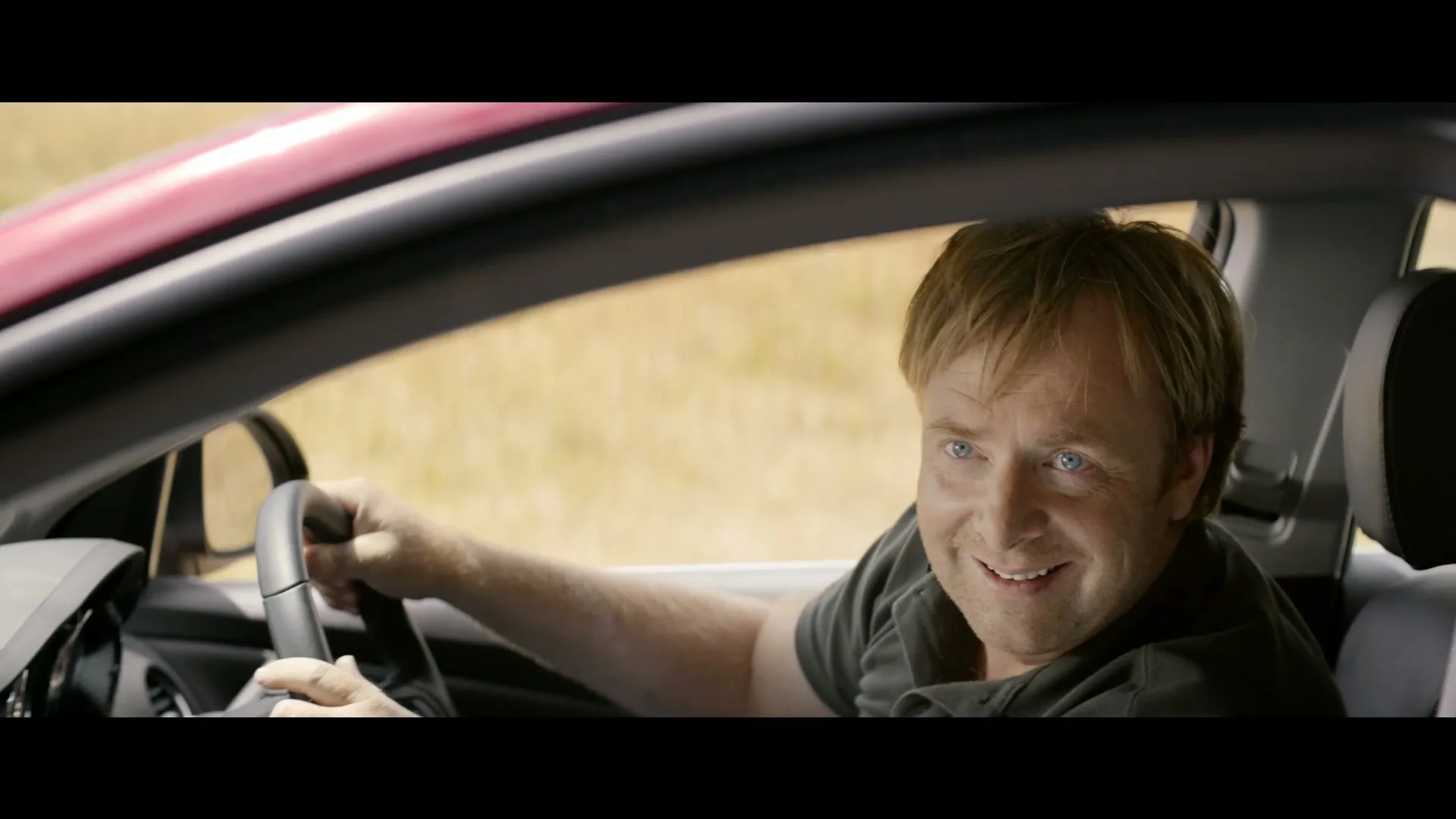 A man with blonde hair and a dark polo shirt smiles while driving a red car on a sunny day.