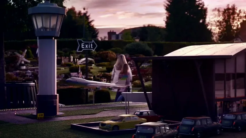 A giant woman in a white coat walks past a miniature airport with tiny cars and buildings, under a pink sky at dusk.