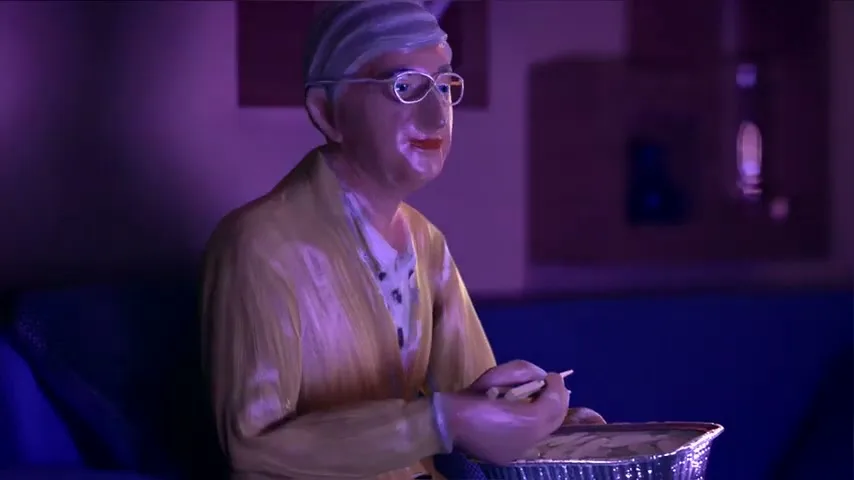 A claymation elderly woman with glasses and a yellow cardigan sits on a blue couch, eating from a foil container in a dimly lit living room.