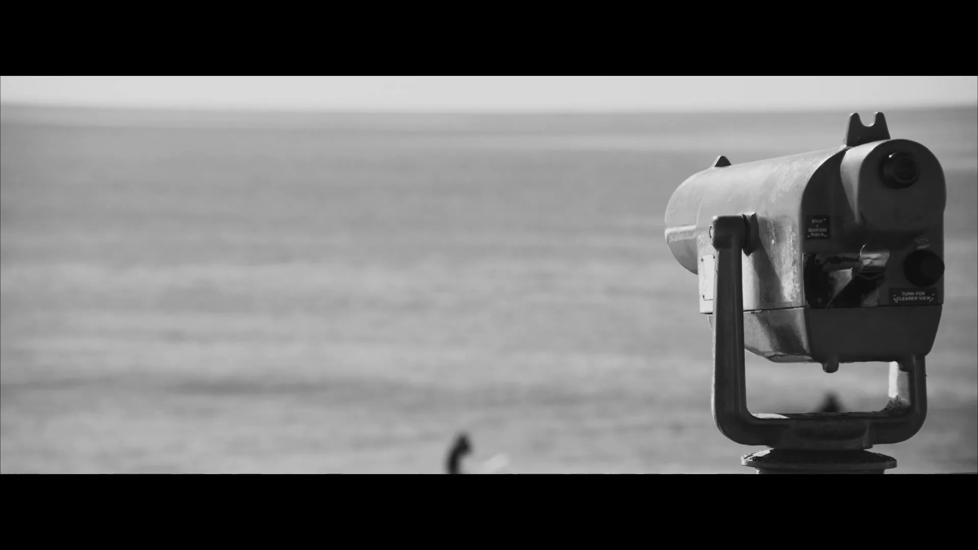 A coin-operated telescope stands on a sunny beach, with a surfer visible in the distant ocean, in a black and white shot.