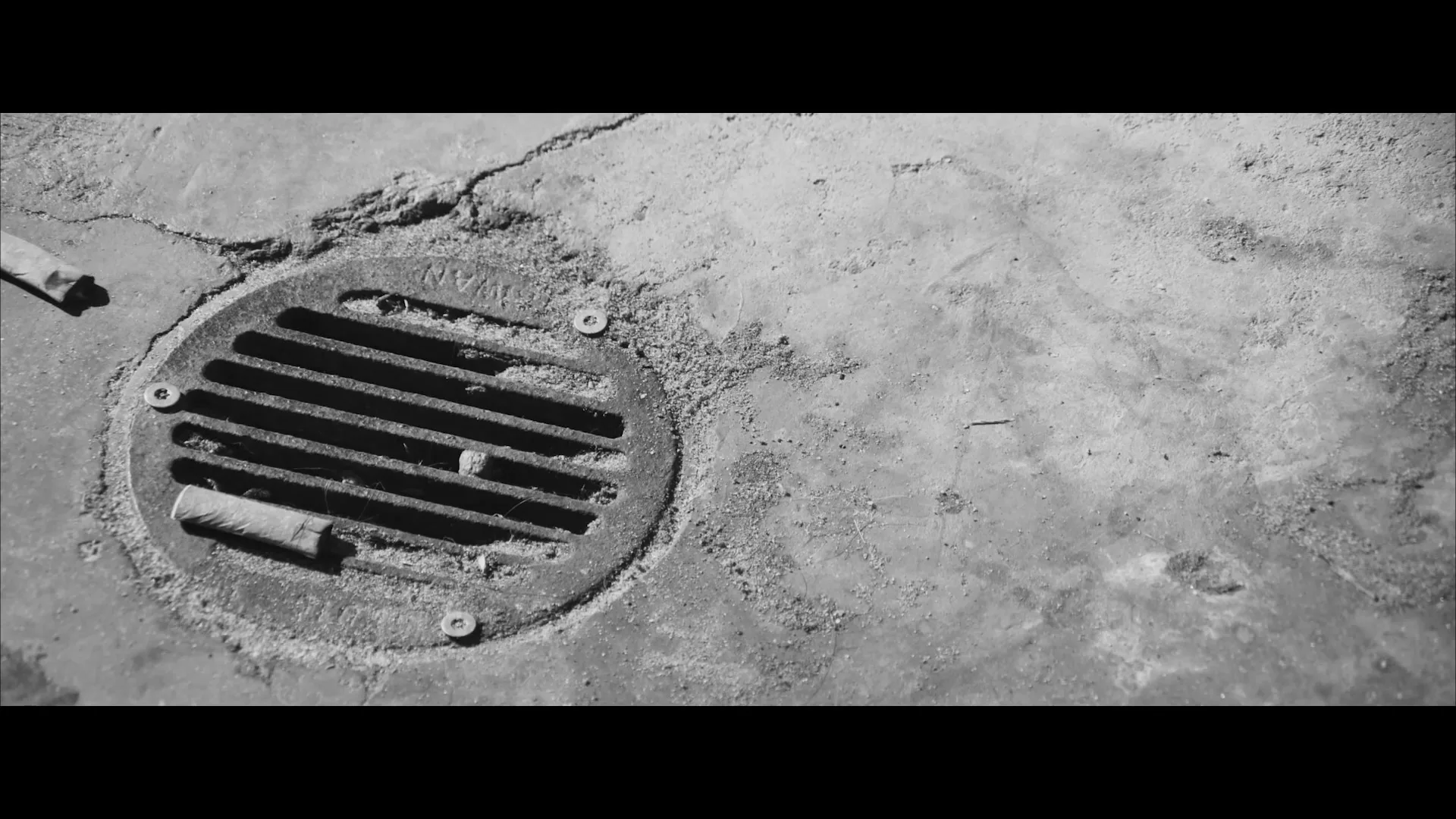 A black and white shot of a coin-operated telescope on a pier, with a surfer in the background, then cuts to a close-up of a dirty storm drain.