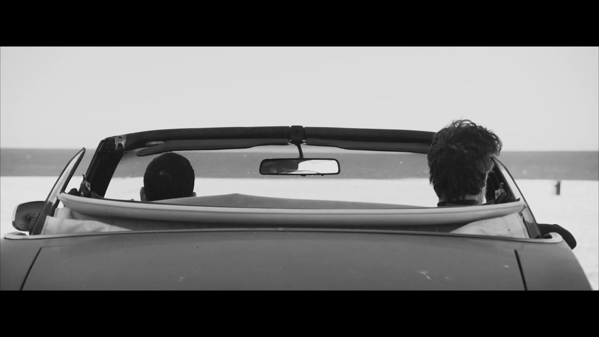 Two men in a convertible car with a surfboard in the back, facing each other and talking, with the ocean in the background on a bright day.
