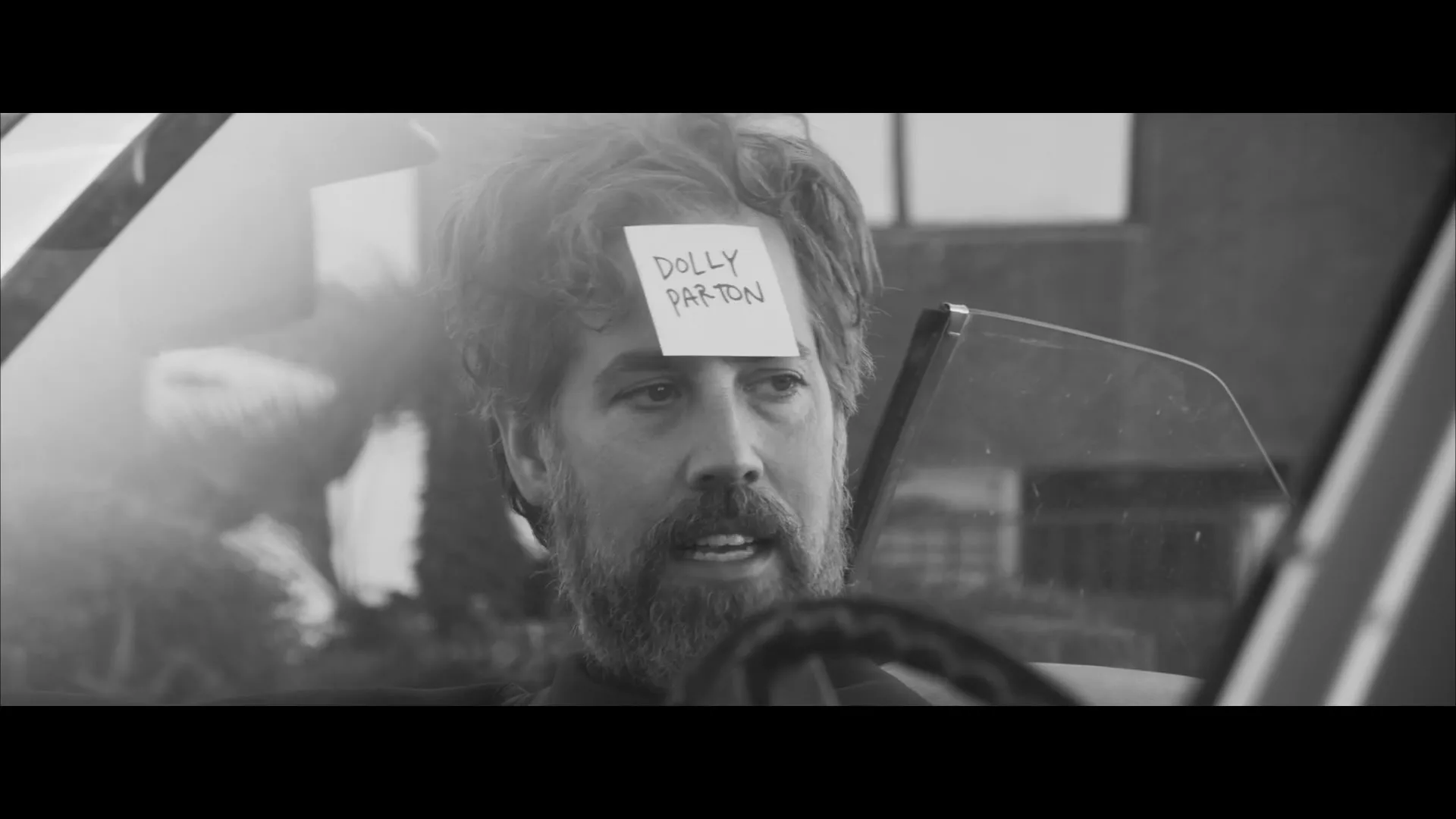 A bearded man with a sticky note on his forehead that reads "Dolly Parton" sits in a car, looking thoughtful and slightly confused.