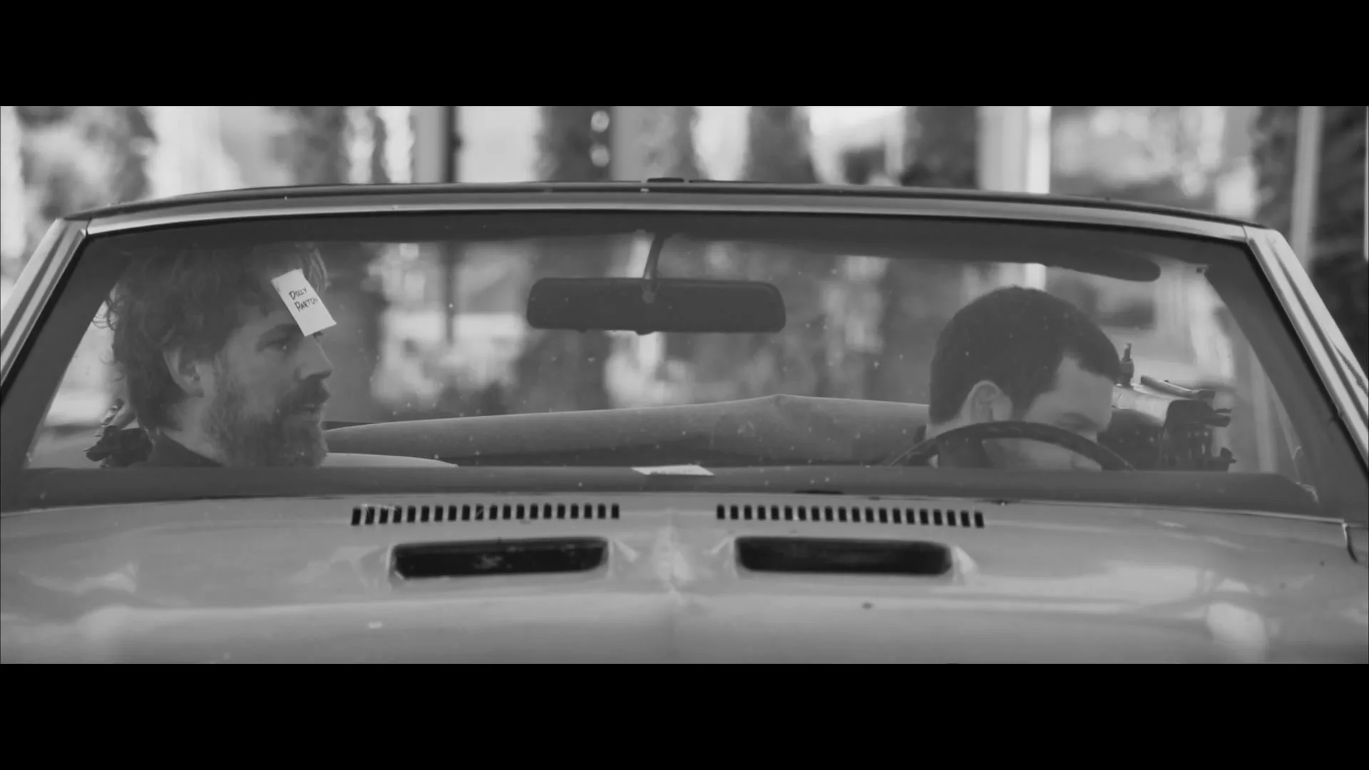 Two bearded men in a convertible car, one driving and the other in the passenger seat, look at each other and talk in a black and white shot.