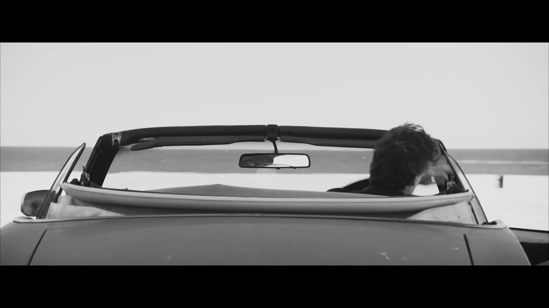 A man with a beard sits in a convertible car with a surfboard in the back, looking out at the ocean on a bright day.