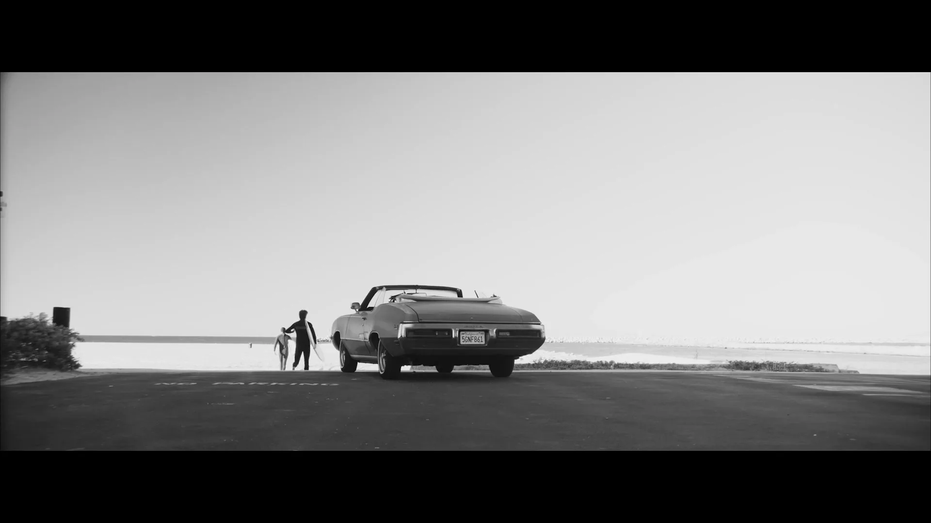 A man in a wetsuit with a surfboard walks away from a classic convertible car towards the ocean on a sunny day.