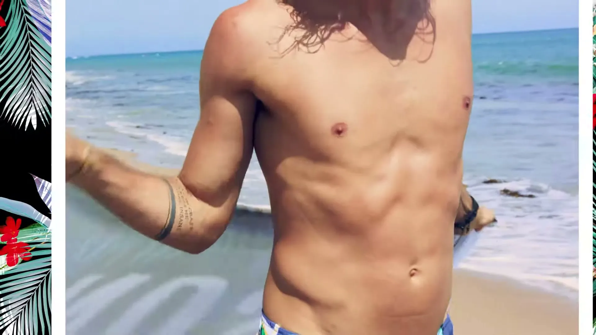 A fit male with tattoos on his arms walks out of the ocean onto a sandy beach, holding a towel.
