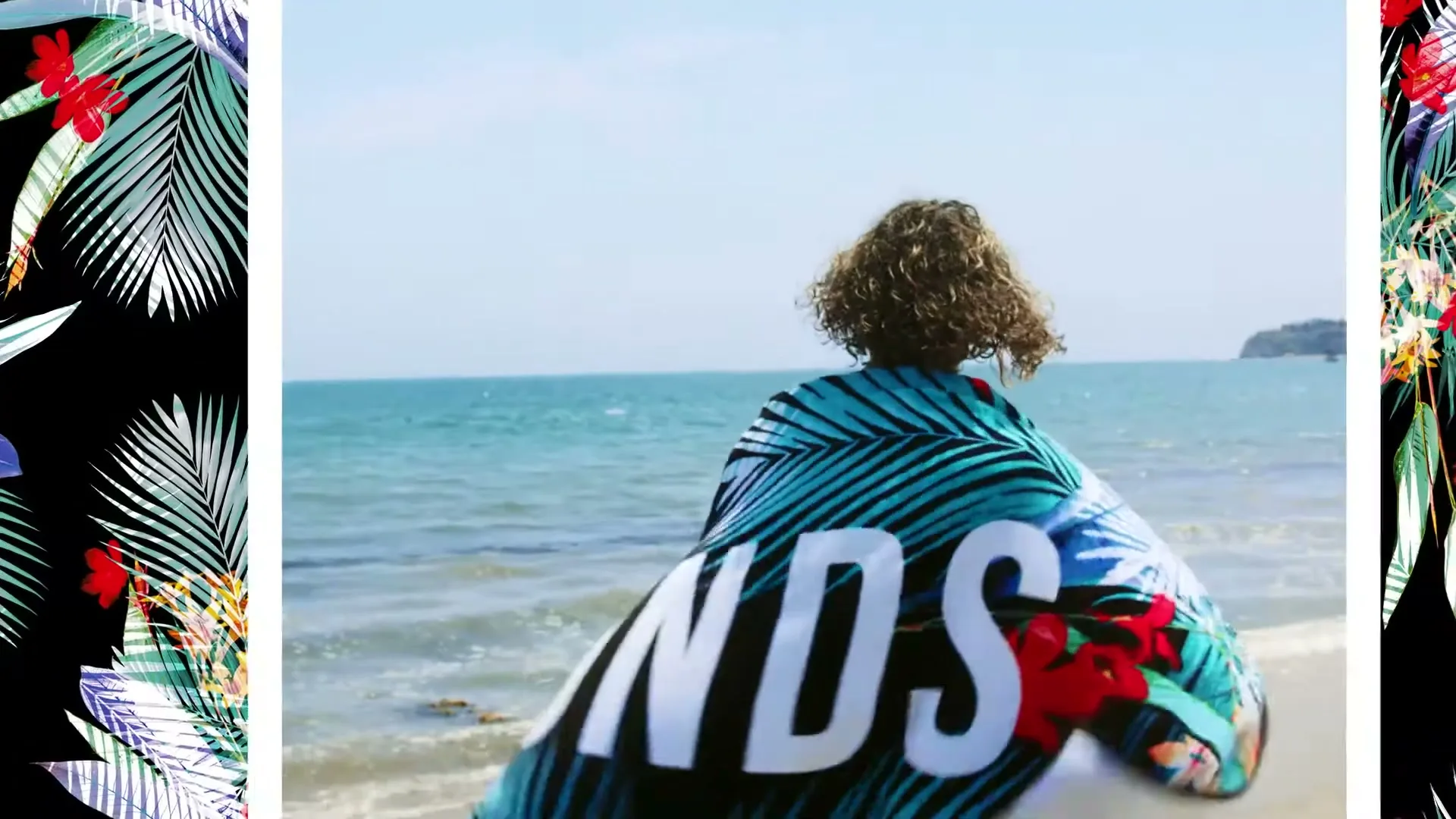 A person with curly hair runs towards the ocean on a sunny beach, wrapped in a blue and black tropical print towel with "BONDS" written on it.