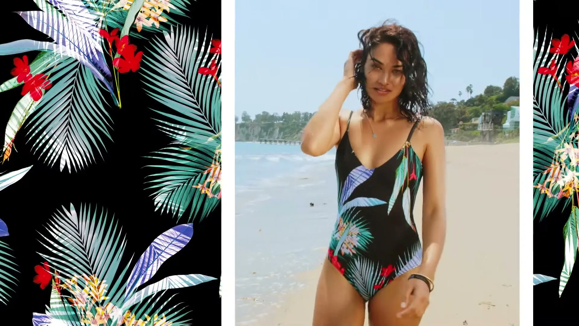 A young woman in a black, tropical print one-piece swimsuit stands on a sandy beach, looking at the camera with a slight smile.