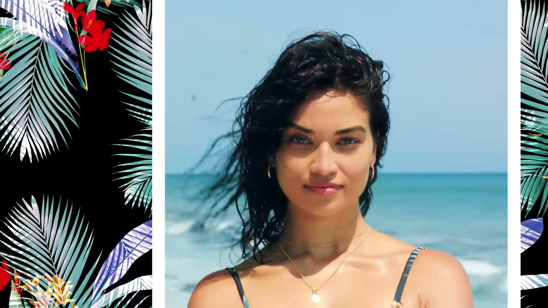 A woman with dark, wet hair and a patterned bikini top smiles at the camera on a sunny beach with ocean waves in the background.