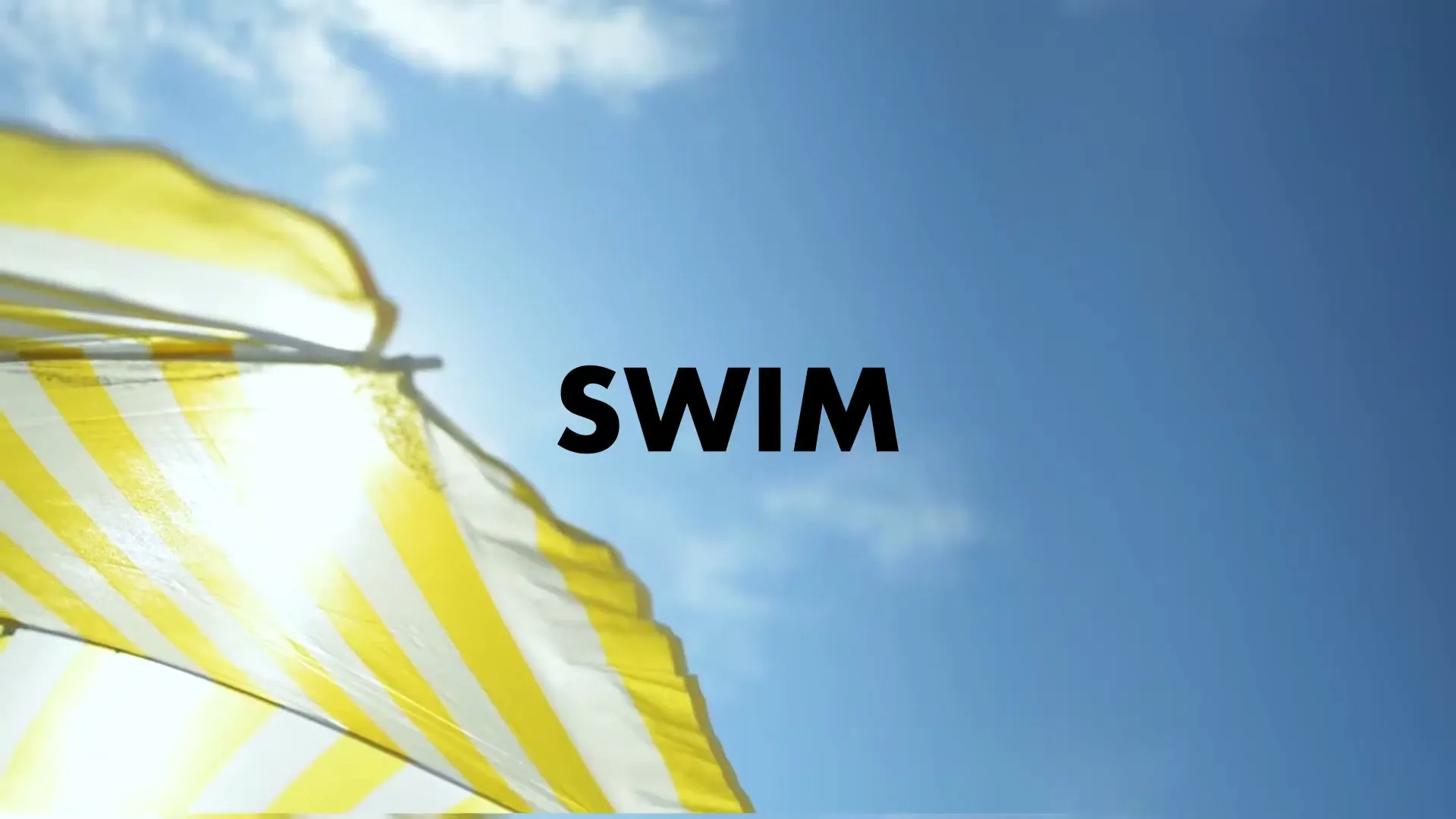 A yellow and white striped beach umbrella is seen from below against a bright blue sky with the word "SWIM" in the center.