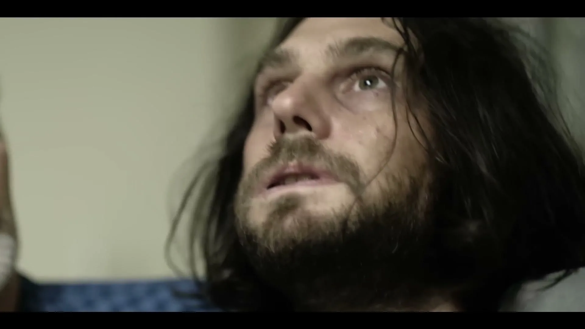 A disheveled man with long dark hair and a beard looks up with a pained expression, possibly in a hospital room.