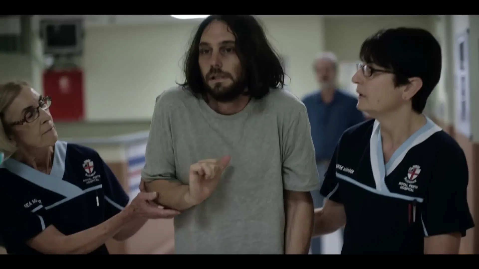 Two female nurses in blue scrubs help a disheveled male patient with long hair walk down a hospital hallway.