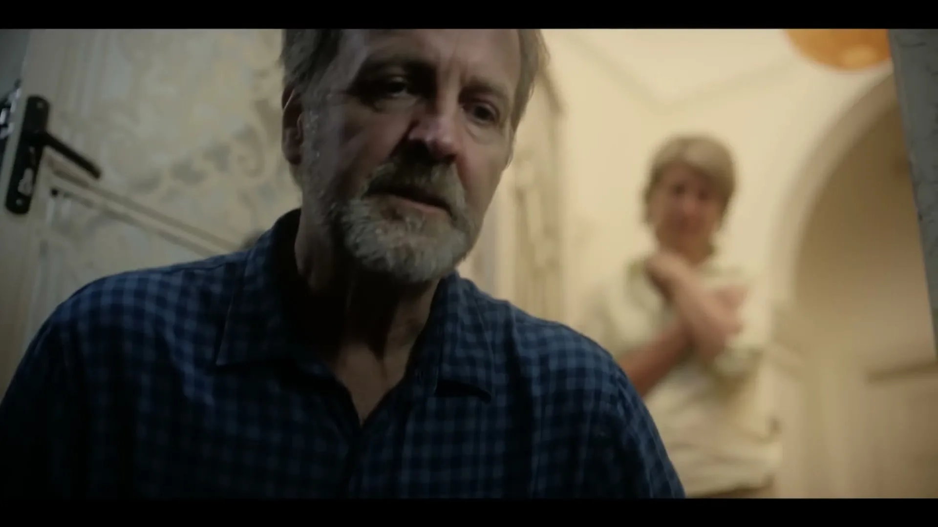 A distressed older man with a beard looks down, while an older woman stands blurred in the background of a hallway.