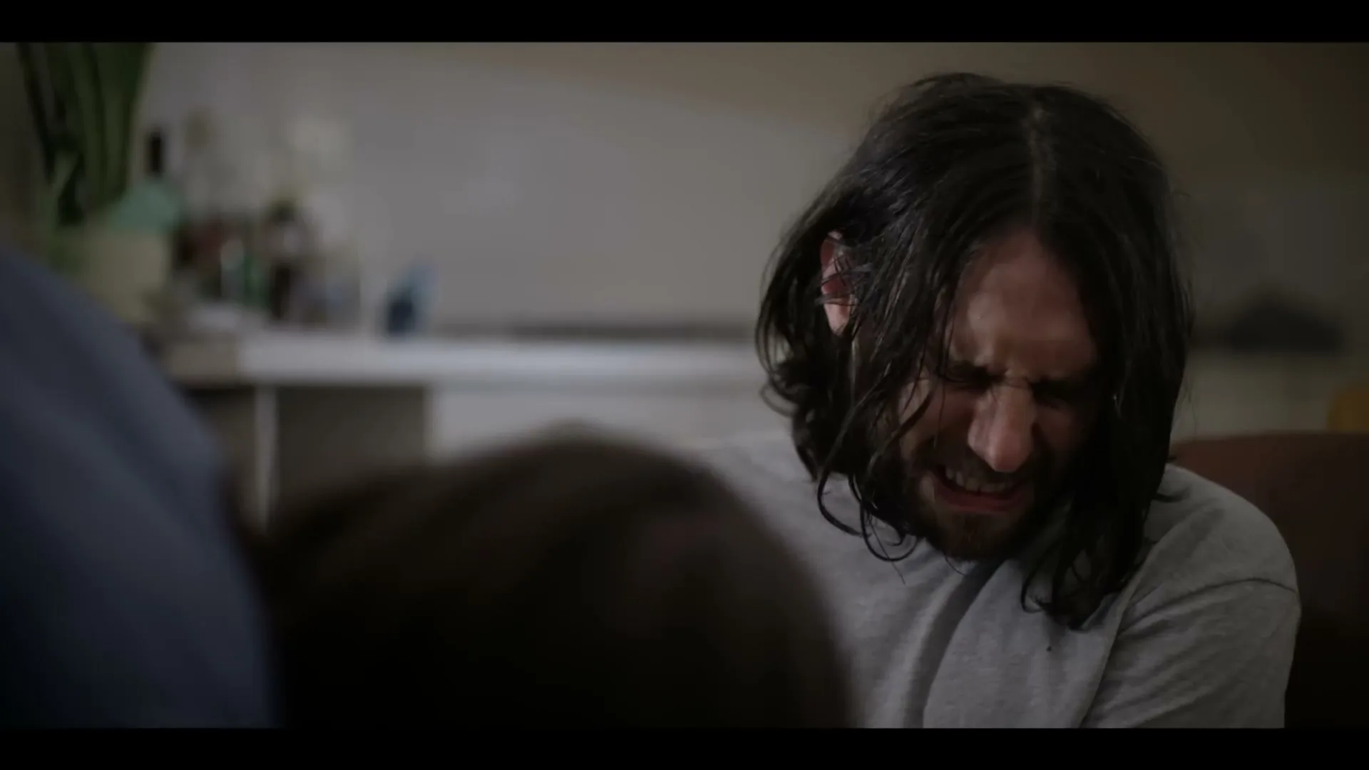 A man with long dark hair and a beard grimaces in pain or discomfort, seen over the shoulder of another person in an interior setting.