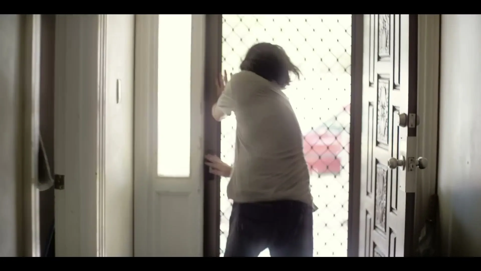 A young man with long dark hair, wearing a t-shirt and jeans, struggles to close a screen door in a home entryway.