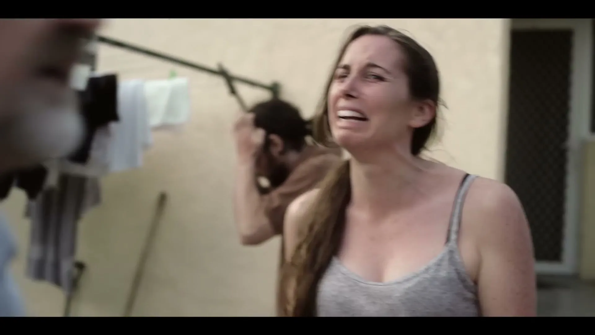 A distressed young woman in a grey tank top screams at an older man, while another man in a brown shirt covers his ears in the background, outdoors.