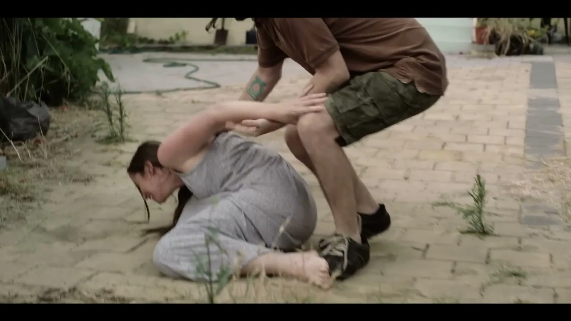 A man in a brown shirt and cargo shorts is physically restraining a woman in a grey dress on a dirt and paved backyard.
