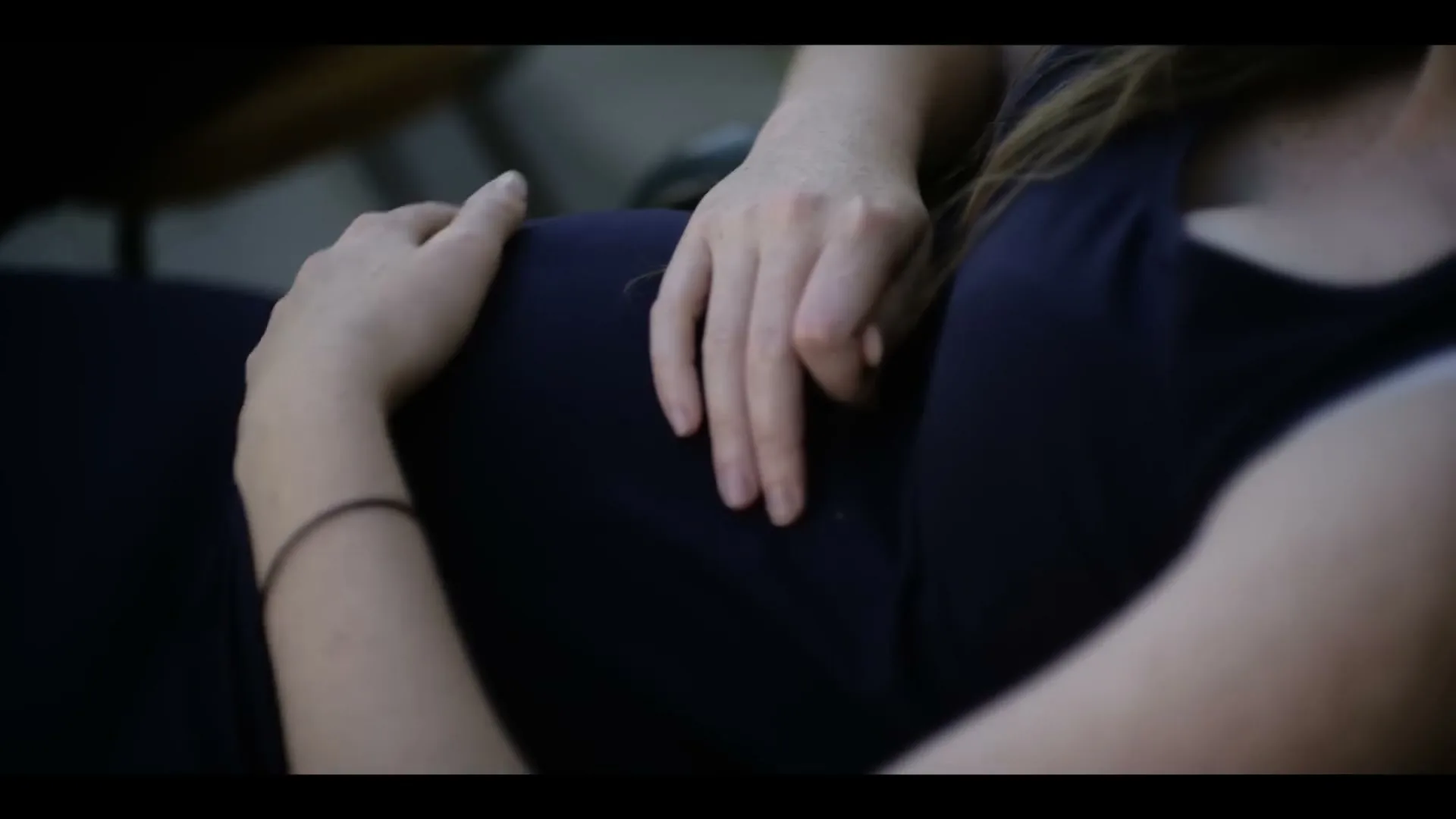 A pregnant woman in a dark blue dress lies down, gently caressing her baby bump with both hands.