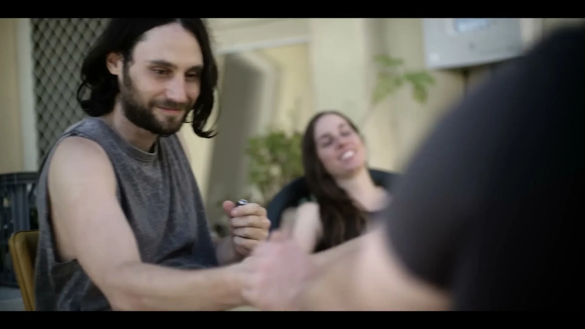 A man with long dark hair and a beard smiles while holding a small object, as a woman laughs in the background at an outdoor gathering.