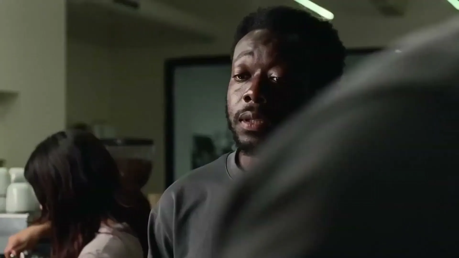 A Black man in his 30s, wearing a grey t-shirt, looks up and smiles at someone off-camera in a dimly lit office kitchen.