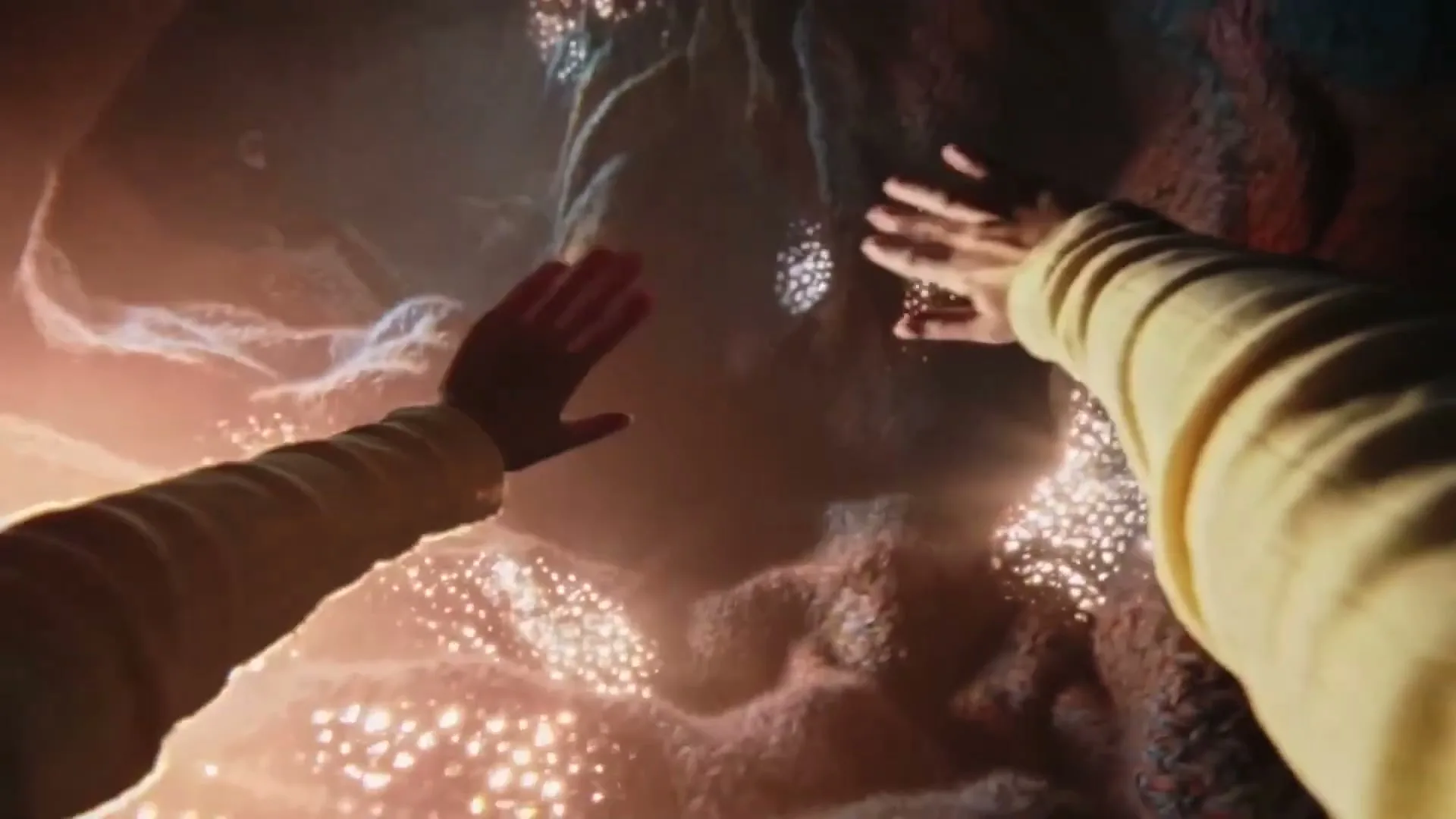 Two people in light-colored long-sleeved shirts reach out their hands to touch a glowing, organic-looking wall in a dark, mysterious cave.