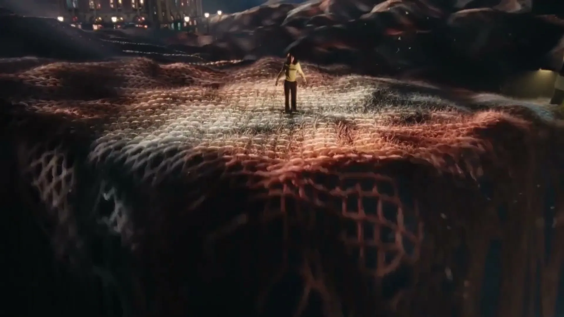 A woman stands on a textured, undulating landscape that resembles a woven fabric, with city lights visible in the distant background under a night sky.